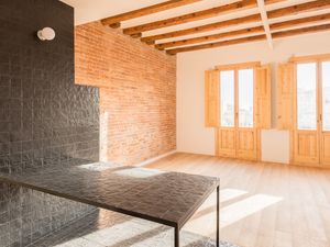 Zona d'estar de pla obert amb encimera de rajoles negres, paret de maó vist, bigues de sostre de fusta i llum natural que entra per dos grans balcons amb persianes de fusta.