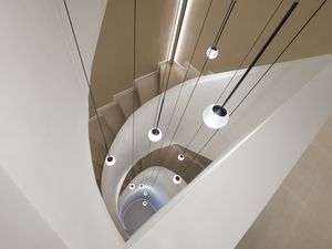 High-angle downward view into the central void of a smooth beige spiral staircase, showing sweeping curved balustrades, integrated recessed lighting along the steps, and a cascade of globe pendant lights suspended on thin black cords.