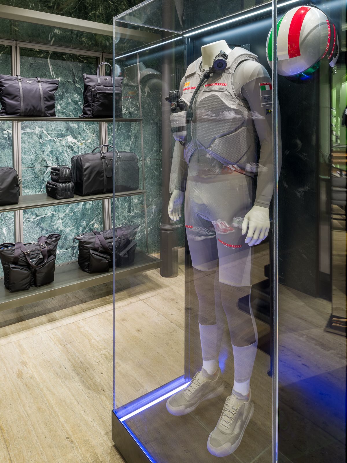 Lujosa boutique Prada con paredes de mármol verde, mostrando un maniquí en un atuendo de rendimiento encerrado en una vitrina de vidrio.
