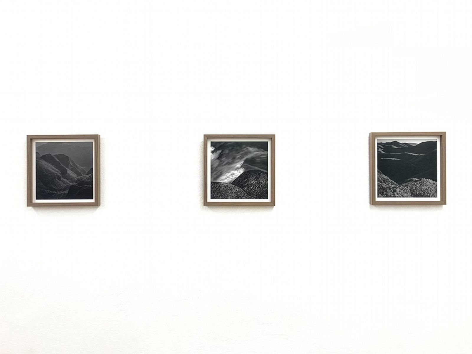 Tres fotografies emmarcades en blanc i negre de paisatges muntanyosos exposades en una paret blanca en un entorn de galeria minimalista.