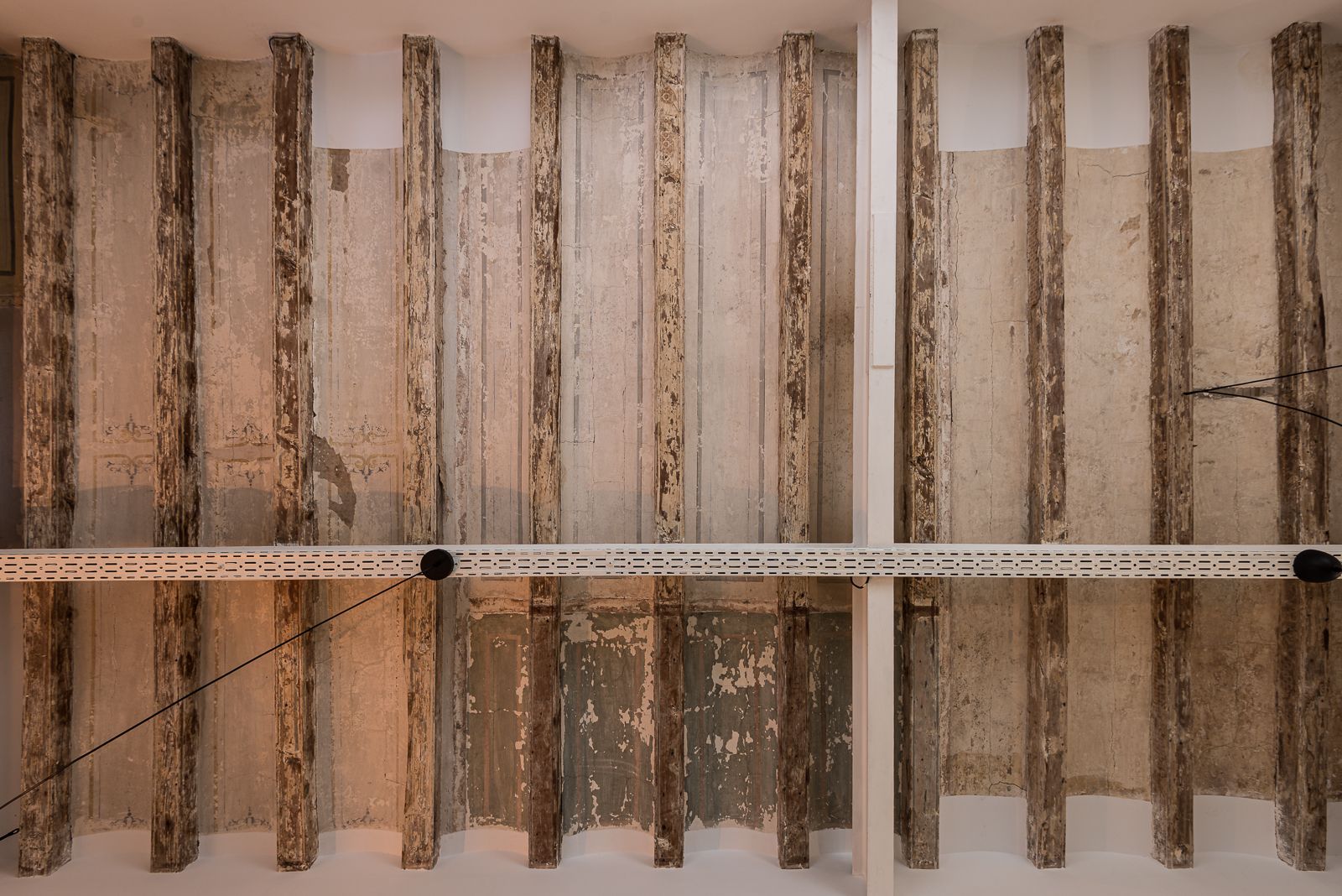 Low-angle shot looking straight up at a traditional Catalan vaulted ceiling featuring exposed weathered wooden beams, distressed plaster with remnants of decorative frescoes, and a modern white cable tray running horizontally across the frame.