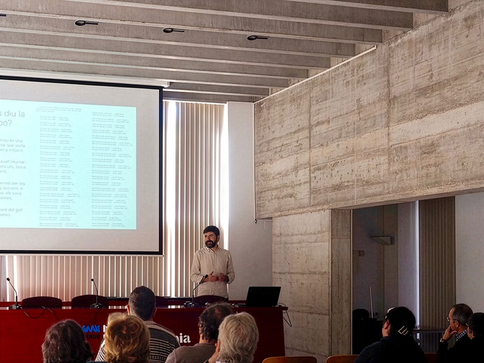 Hombre dando una presentación en una sala de conferencias con una pantalla proyectada y audiencia sentada.
