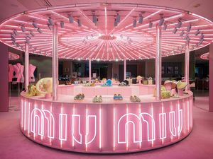 Exposició de botiga emergent Miu Miu il·luminada en rosa amb bosses, sabates i barrets sota un dossel de llums roses brillants.