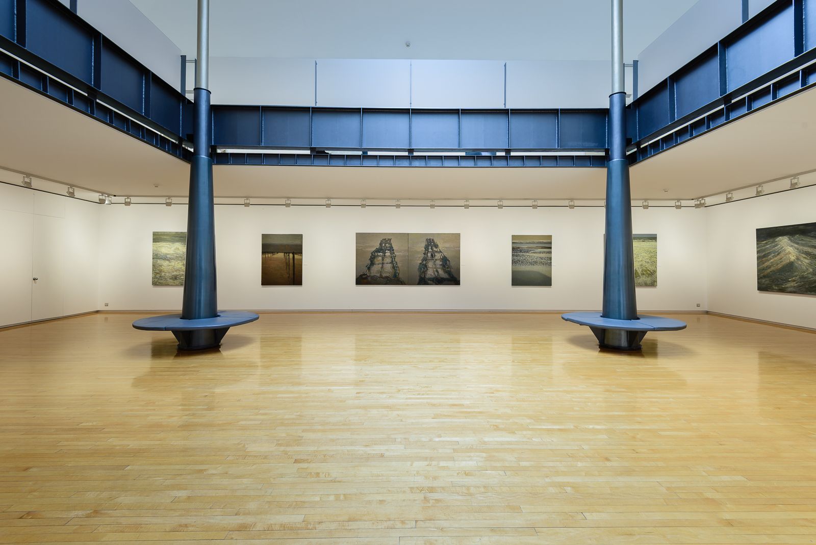 Una espaciosa galería de arte con suelos de madera pulida, paredes blancas y pinturas de paisajes marinos. Dos columnas negras con bancos circulares se encuentran al lado de un atrio cubierto con tragaluz.