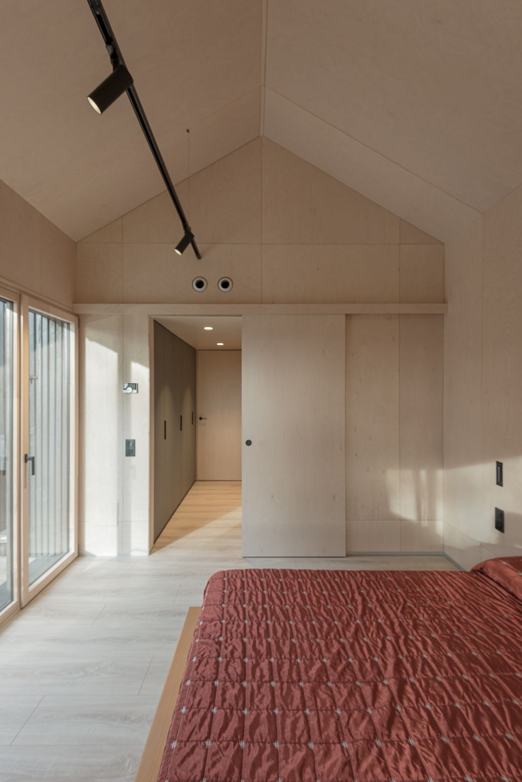 Dormitorio con un techo alto inclinado, con una colcha roja, grandes puertas de vidrio y paredes y suelos de madera minimalistas.