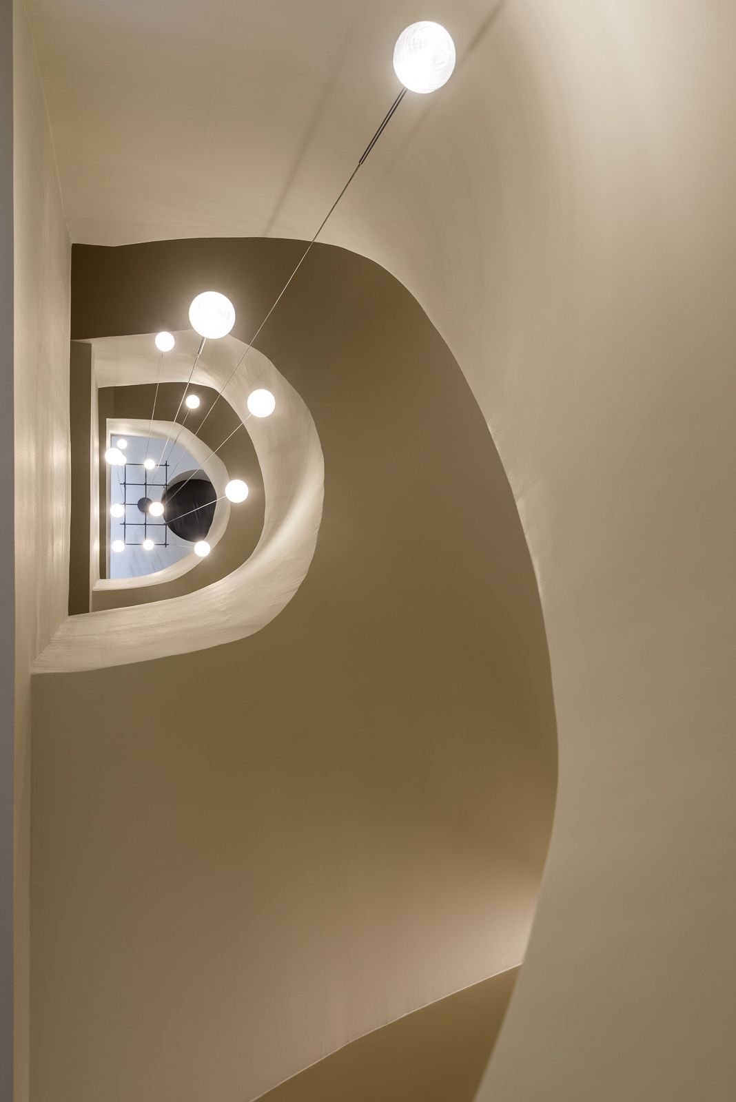 Low-angle view looking upward through the central void of a smooth, beige spiral staircase, framed by sweeping architectural curves and suspended glowing orb pendant lights descending through the multi-story opening.