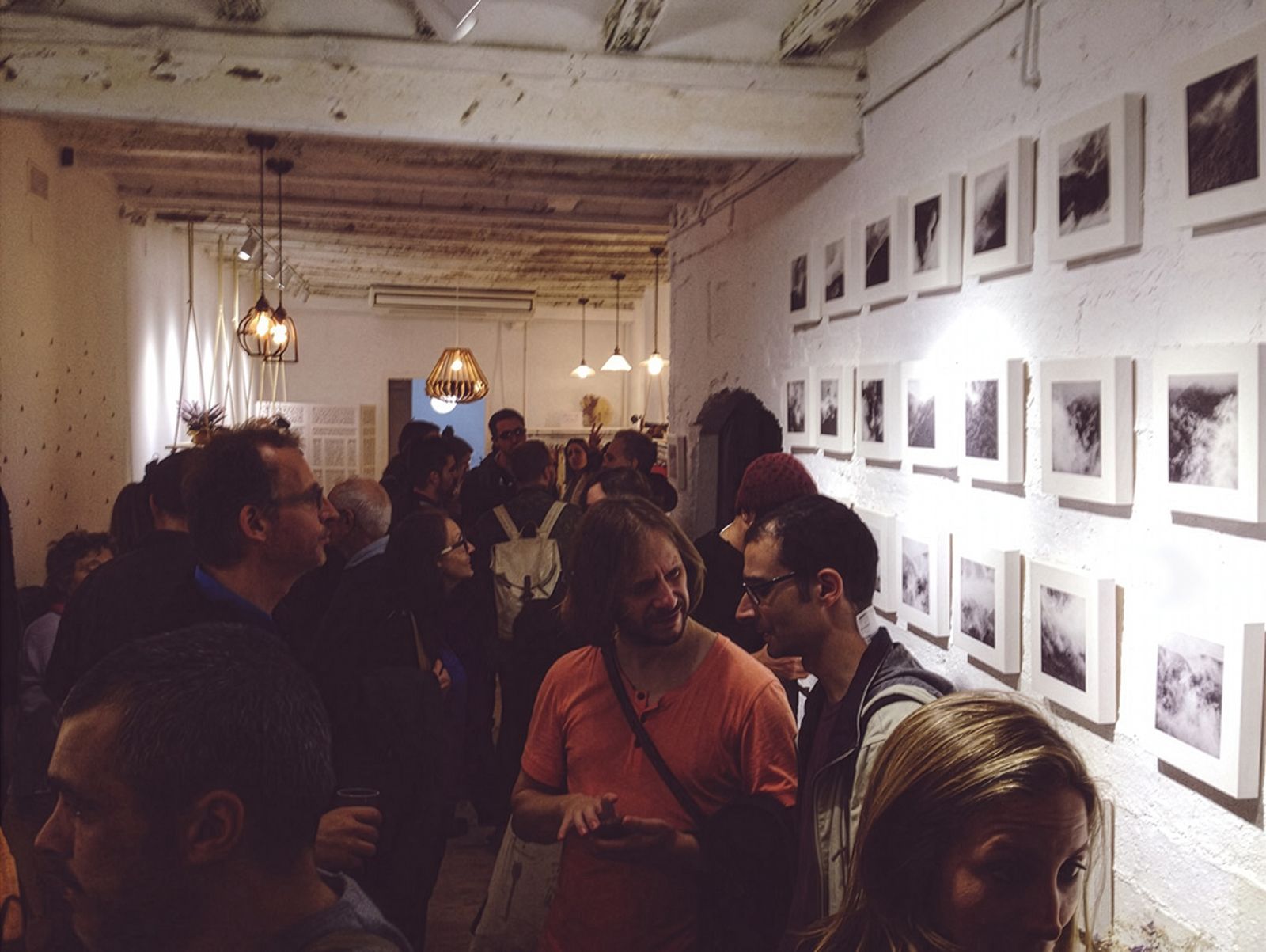A crowded art gallery with people viewing black and white photographs of clouds and mist arranged in a grid on a white brick wall.